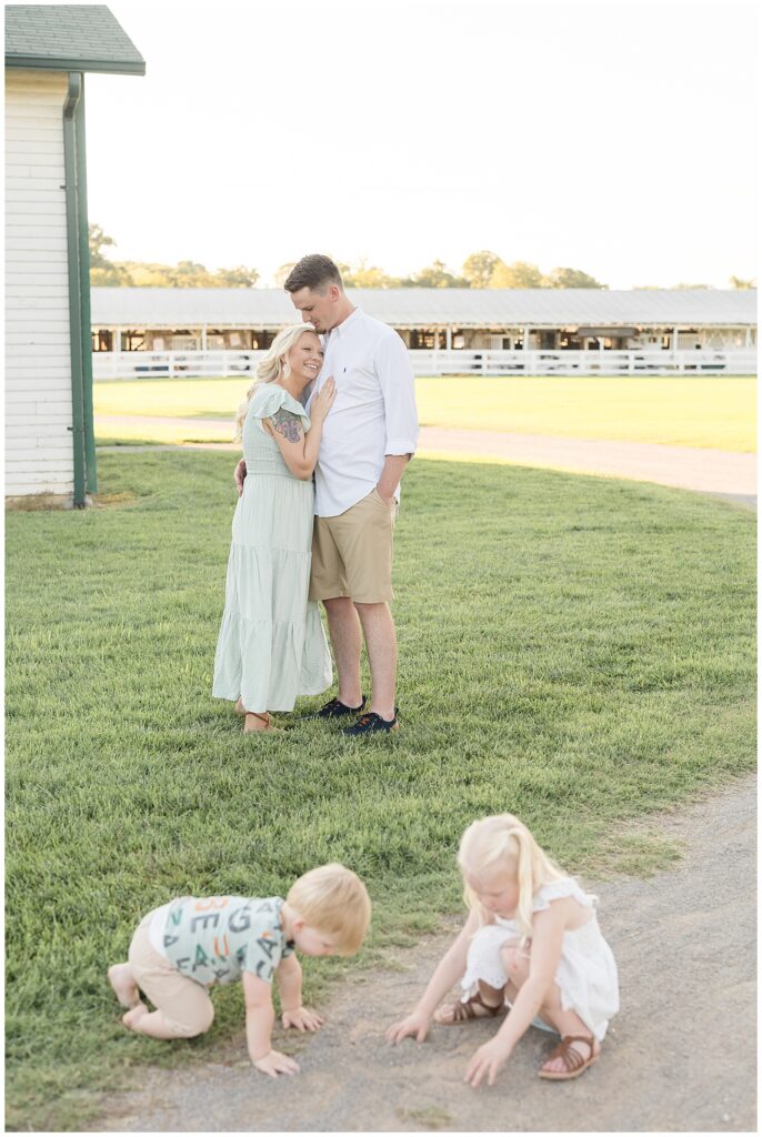Couple stands in the background as they share a hug as she rests her head on his chest and their two young children play in front of them in the gravel during their family portraits.  Click to see more on the blog today!