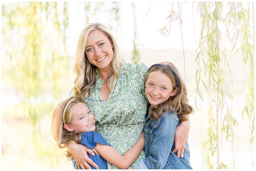 Mom wears a green and blue floral, v-neck dress as she puts an around around both of her daughters as the three all smile during their family session at Nickajack Lake!