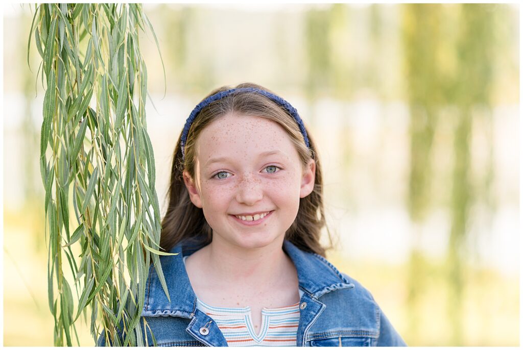 Young girl wears a rainbow striped shirt, jean jacket, and a blue headband as she stands near a willow tree and smiles at the camera of Wisp + Willow Photography Co. with her blue eyes and freckles!