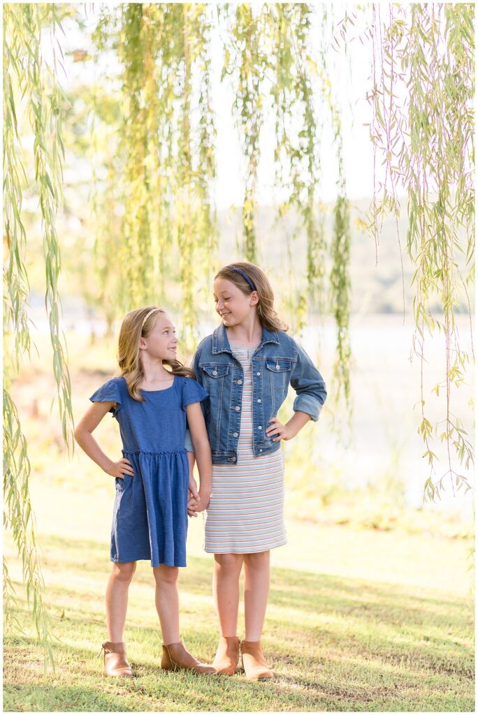 Two sisters hold hands as they look at each other with their other hand on their hip as they wear causal dresses during their family mini session at Nickajack Lake in Chattanooga, TN.