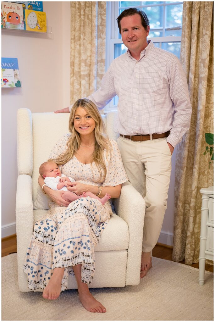 New Mom sits in a rocking chair in her daugher's nursery as she holds her baby girl and smiles at the camera of Wisp + Willow Photography Co.  New Dad stands to the side of the chair with his arm on the back and smiles at the camera for their new family portraits as a family of 3.