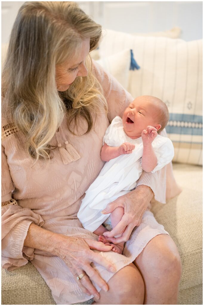 New Grandma, wearing a blush colored dress, holds her new granddaughter cradled in her arms as baby girl smiles big looking up at her!