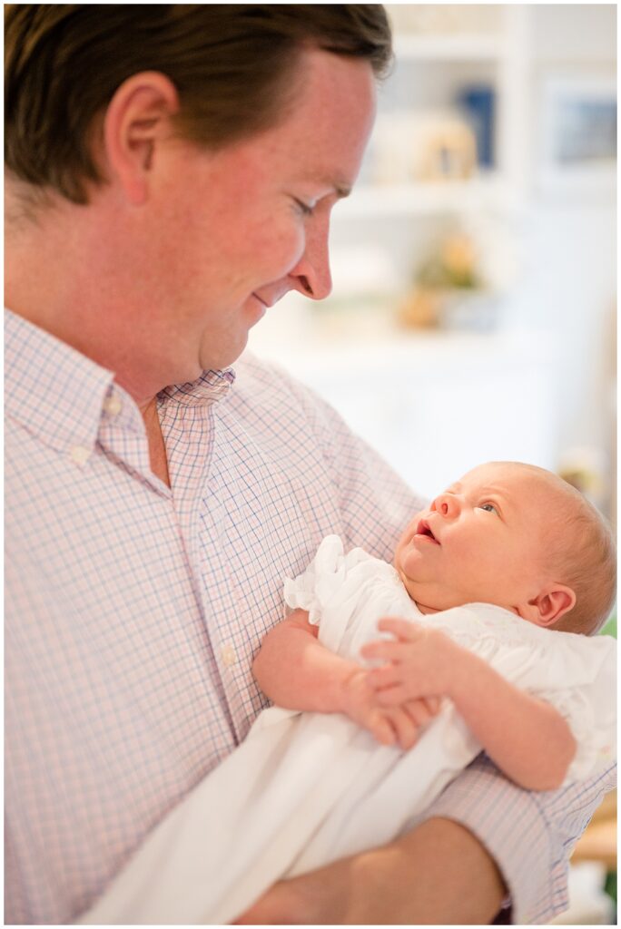 New Dad holds his baby girl cradled in his arms as he looks down at his daughter, wearing a white dress and looks up and smiles at her Dad.