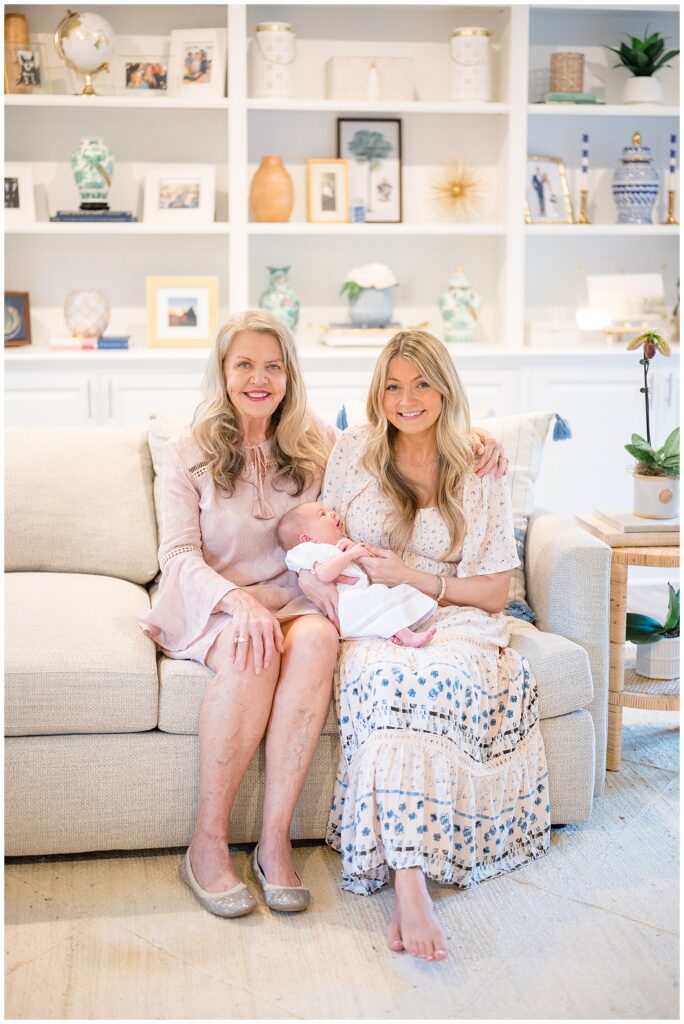 3 generations consisting of baby girl, new mom, and grandma sit on a light grey couch in the coziness of their home as they smile at the camera and baby girl looks up at her mom.