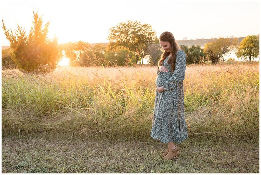 Mom-to-be wears a long, green dress with small white flowers as she stands in a field in front of the White Rock Lake in Dallas, TX.   She holds her belly and looks down.