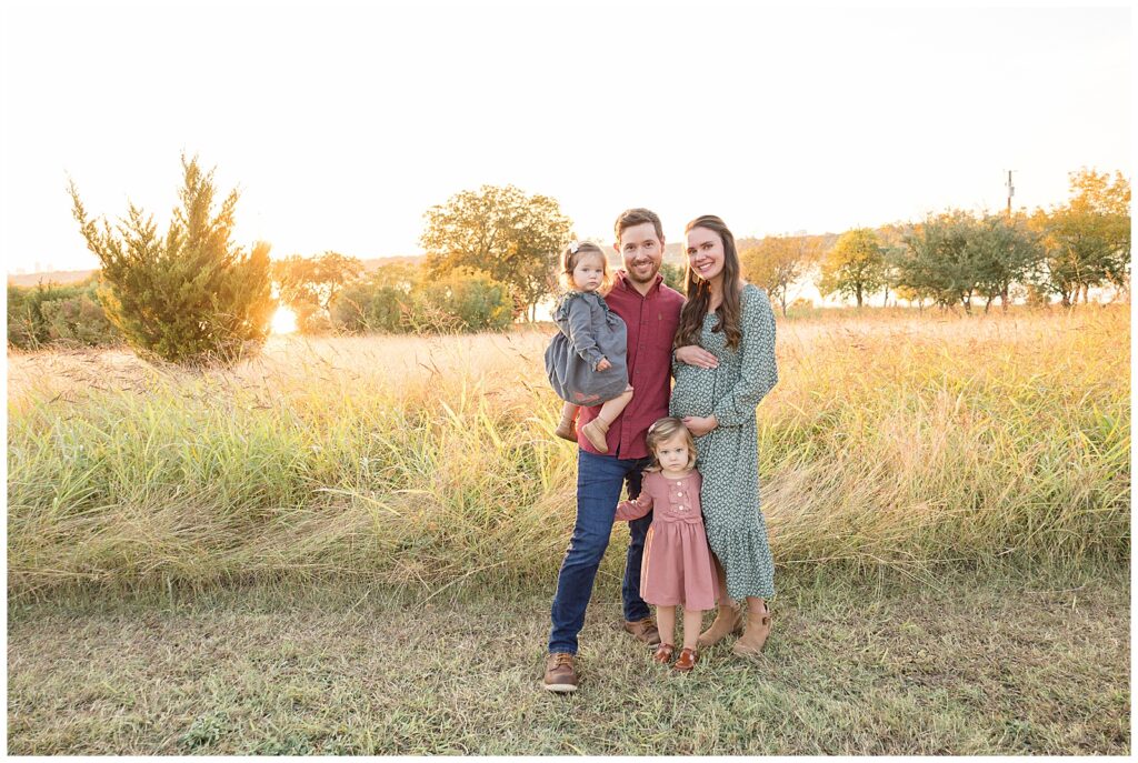 Family of 4 and expecting a new baby, stand in field in front of White Rock Lake as Dad holds youngest daughter, mom holds her belly, and their toddler daughter stands and hugs both Mom and Dad's legs.