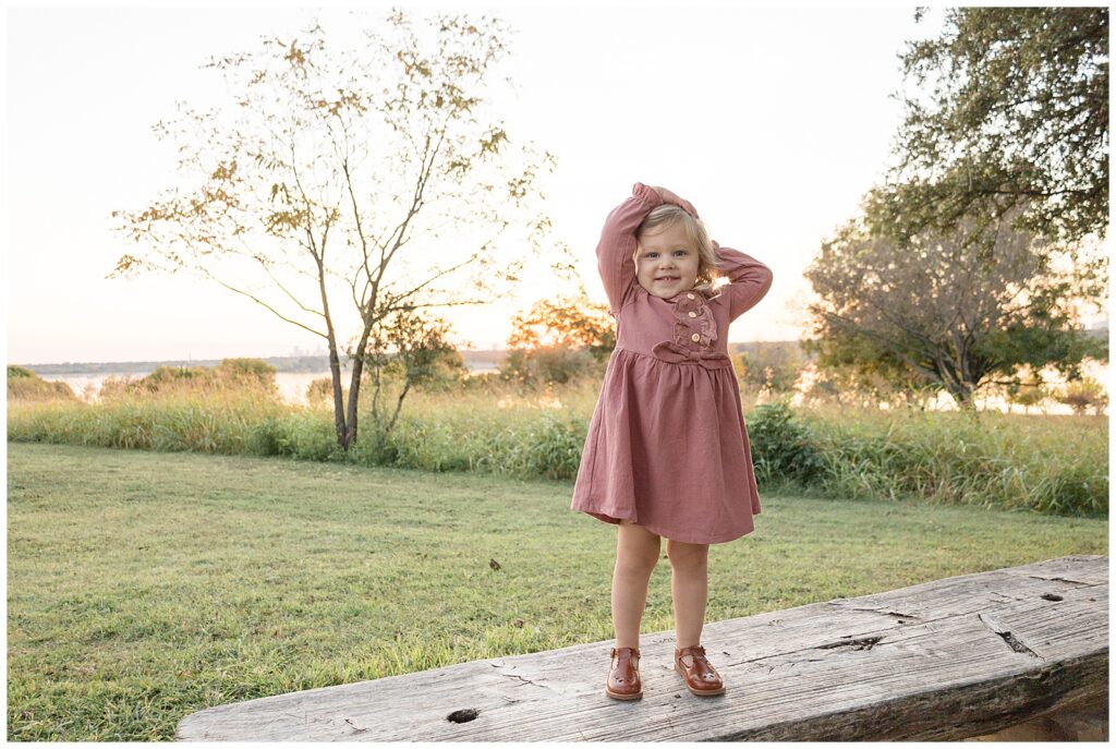 Toddler girl wears a dark pink dress with Mary Jane brown shoes and holds her head as she smiles at the camera of Wisp + Willow Photography Co. in Dallas, TX.