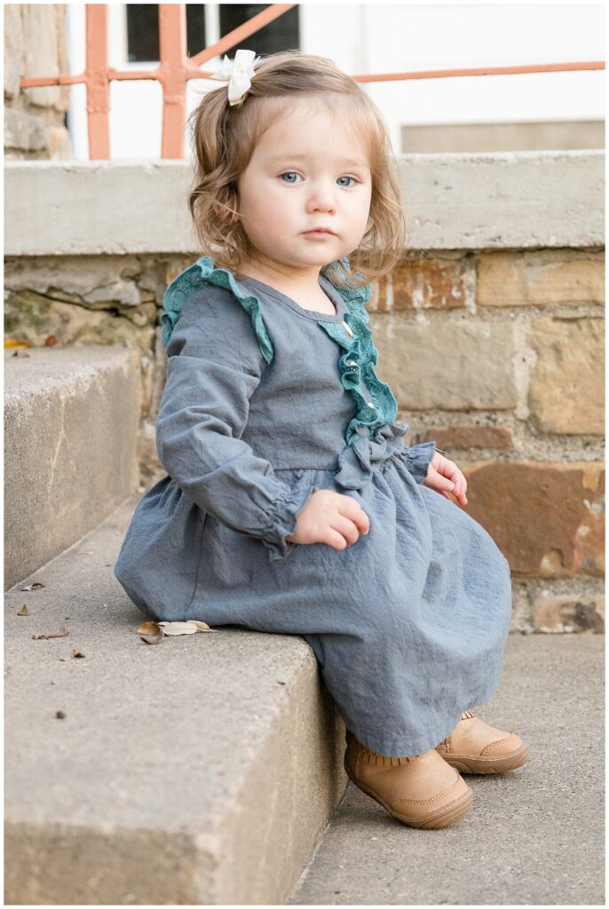 Little girl wears a denim dress with teal fringe on the sleeves and down the front with the buttons and a white bow pulling her hair back as she sits on concrete steps and looks at the camera of Wisp + Willow Photography Co.