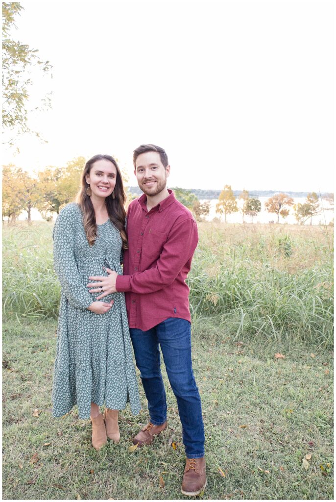 Couple stands in field in front of White Rock Lake during their Dallas portrait session as husband holds his wife's belly as they prepare for baby #3.