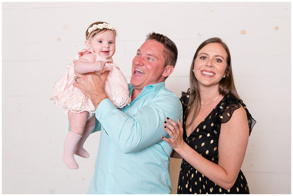 Family of 3 take family portraits in front of white cylinder wall.  Mom wears a black v-neck dress with gold dots and holds on to her husband from behind who is holding their baby girl who is wearing a pink formal dress and a floral headband.