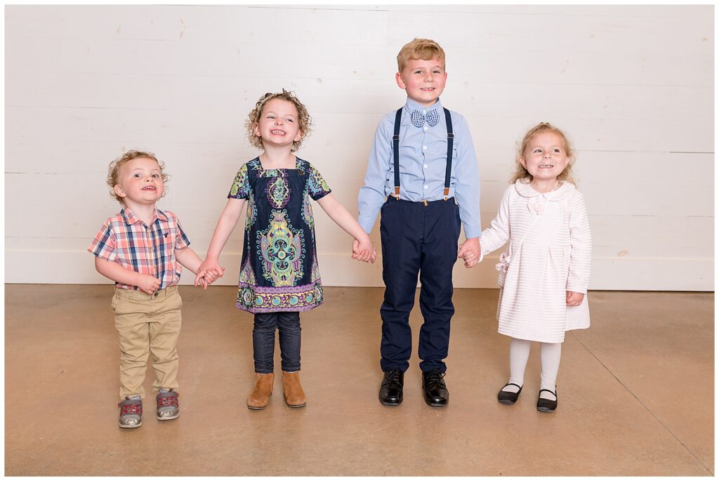 4 cousins hold hands as they stand together all dressed up with dresses, a bow tie, and a button down.  They smile at the camera of Wisp + Willow Photography Co. who is capturing extended family portraits.
