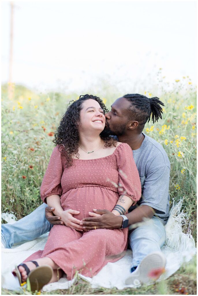This maternity session in Plano, TX has pregnant wife sitting in a wildflower field leaning back in her husbands lap as he kisses her on the check.  Click to see more of this session from Wisp + Willow Photography Co. on the blog!