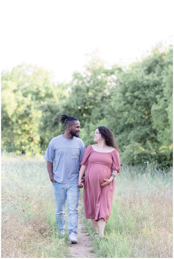 Couple walks down a path surrounded by wildflowers holding hands and looking at each other as expecting momma holds her belly!
