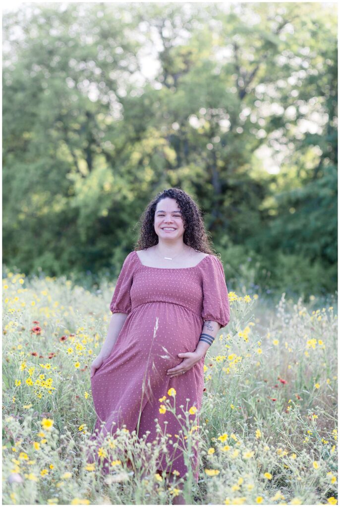 Plano, TX maternity session at Arbor Hills Nature Preserve has pregnant momma standing in a wildflower field, smiling at the camera and holding her mauve colored dress with one hand and her belly with the other.