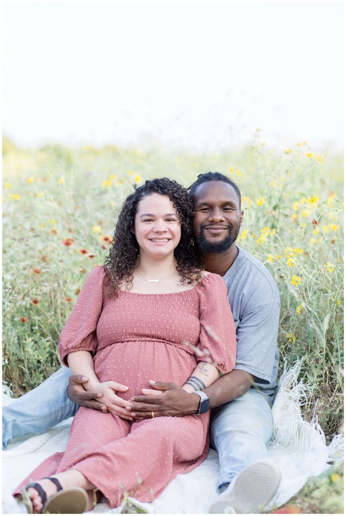 Plano maternity session has couple sitting in a field surrounded by wildflowers as the husband sits behind his wife and they both smile cheek to cheek at the camera while holding the belly of their baby on the way!  Click to see more of this couple on the blog today!