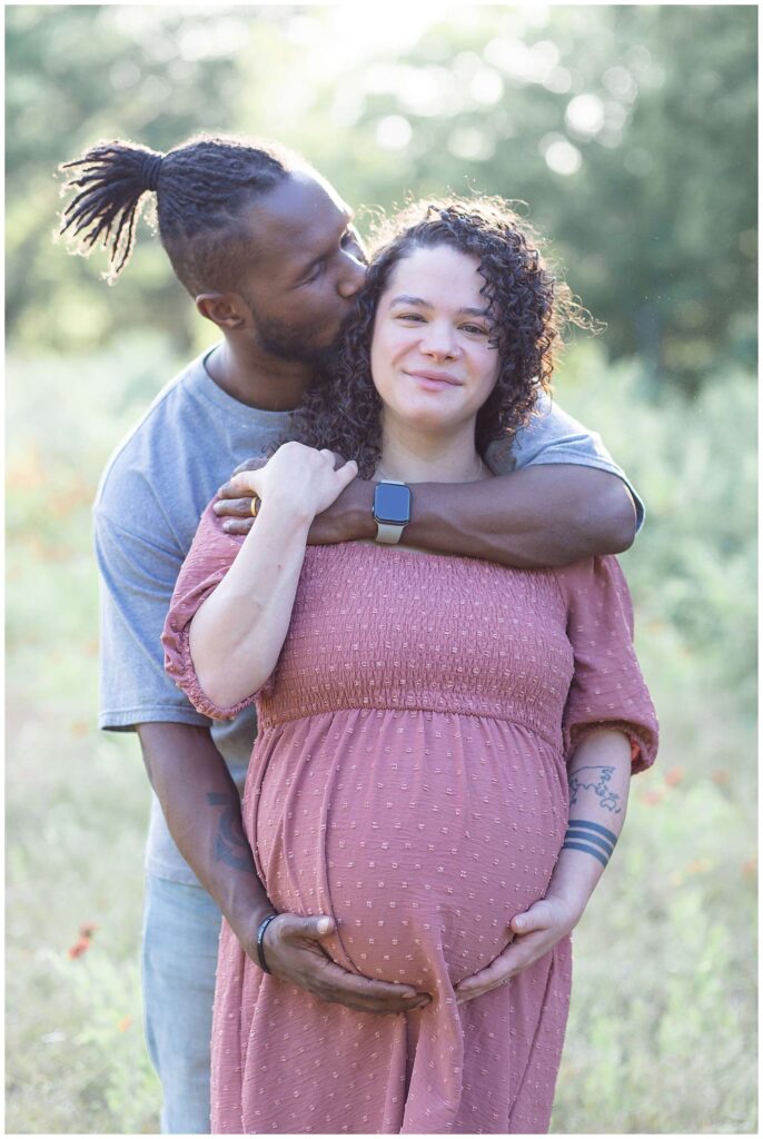 Couple takes maternity portraits at Arbor Hills Nature Preserve in Plano, TX.  Husband stands behind his wife, holding her belly and giving her a bear hug while expecting momma holds his hand and holds her belly while smiling at the camera of Wisp + Willow Photography Co.