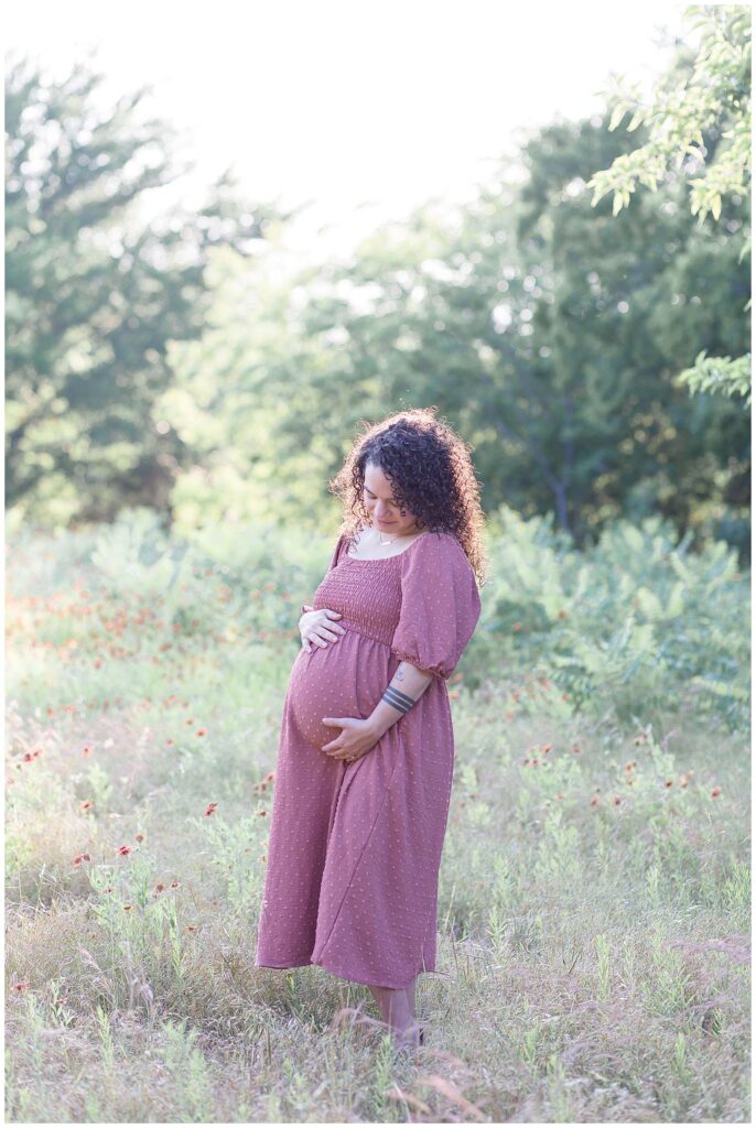 Pregnant momma stands in a field surrounded by wildflowers wearing a long raspberry colored dress for her maternity session in Plano, TX.