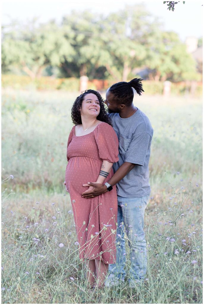 Expecting couple stand in a field for their Plano maternity session as they hold the belly of their baby on the way and the wife turns back to look at her husband as they smile at each other!