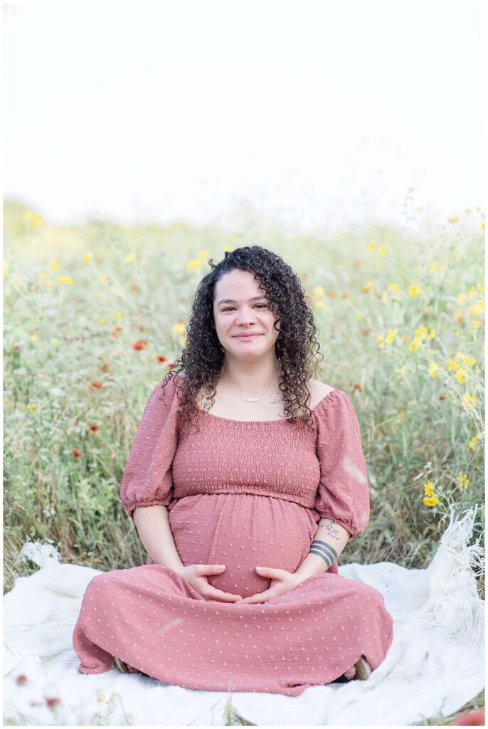 Pregnant momma sits in a field of wildflowers in Plano, TX wearing a mauve colored dress, holding her belly and smiles at the camera of Wisp + Willow Photography Co. during her maternity session.  Click to see more from this session on the blog!