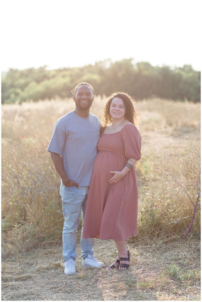 Couple stands in a field in Plano, TX for their maternity session.  Wife wears a long, mauve colored dress and her husband wears a grey t-shirt and jeans.  They stand next to each other as pregnant momma-to-be holds her belly and they smile at the camera of Wisp + Willow Photography Co.