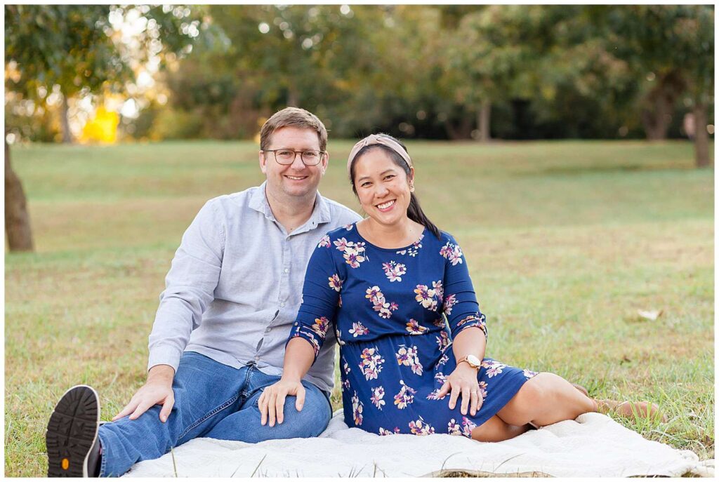 Parents take a minute to take a picture of just the two of them during their Raleigh family portrait session. Couple sits on the grass together as they smile for Wisp + Willow Photography Co.  Wife wears a blue, floral dress with a neutral headband and her husband wears a denim shirt and jeans.