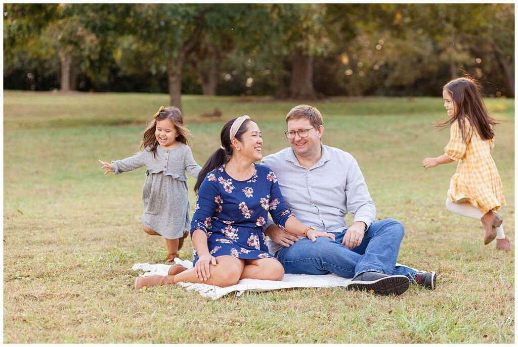 Mom and Dad sit on the grass on a blanket and laugh with each other as their two, young daughters run around them laughing.  Mom wears a blue, floral dress, Dad wears a grey, button-down shirt and jeans, one daughter wear a grey, waffle dress and the other wears a yellow and plaid dress.