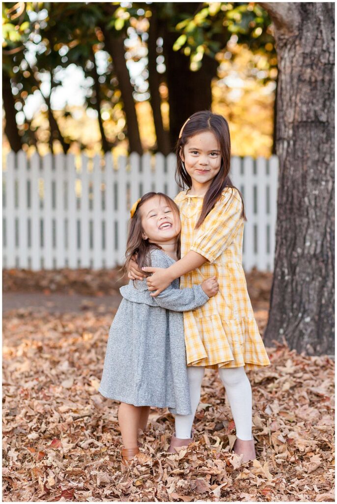 Two sisters stand and give each other a hug while smiling at the camera of Wisp + Willow Photography Co. during their fall portrait session in Raleigh, NC.  They coordinate wearing yellow and grey colors as they stand in the fall leaves in front of a white, picket fence.