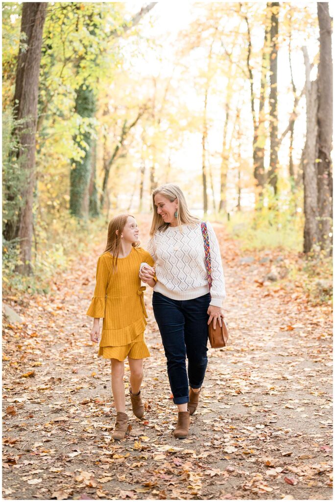 A mom and daughter walk hand in and hand down a tree-lined path covered with leaves at Belle Isle in Richmond, VA during their fall family portrait session.