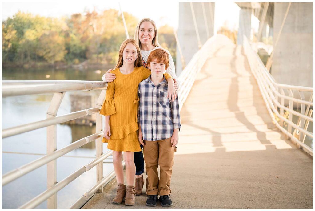 Mom stands behind her red-headed son and daughter with her hands on their shoulders as they stand on the bridge at Belle Isle in Richmond, VA.