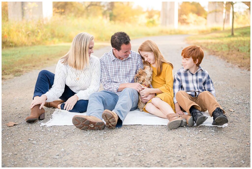 Family of four, wearing coordinating colors of fall yellow, white, and blue, sit on a blanket on a pathway as they look at their little pup.  See more of this family on the blog today!