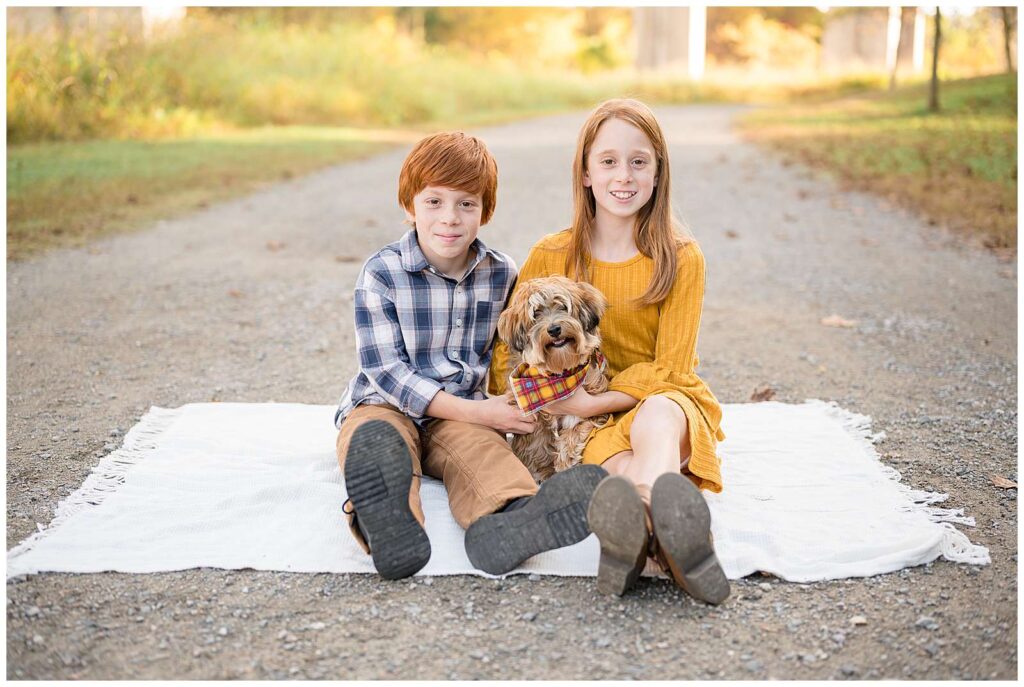Two red-headed siblings, one sister and one brother, sit on a blanket with their little pup in between them as they all look at the camera of Wisp + Willow Photography Co.
