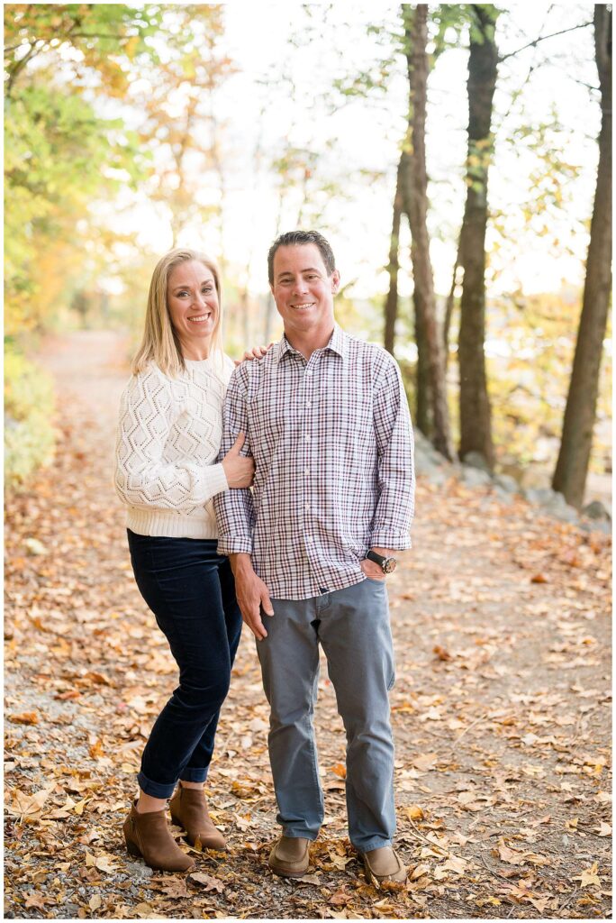 Couple stand on a leaf covered path at Belle Isle in Richmond, VA and smile at the camera of Wisp + Willow Photography Co.  Wife holds on to her husbands arm and shoulder and he puts one hand in his pocket and the other down by his side.