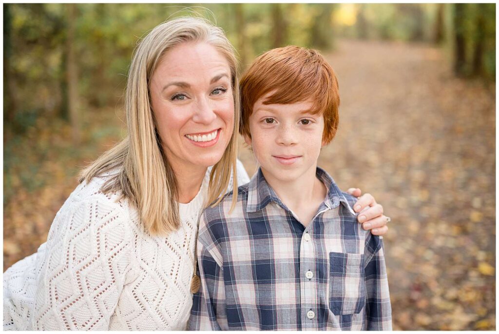 Mom bends down and puts her arm around her son's shoulder.  She wears a white textured sweater and her son wears a plaid shirt during their fall family session.