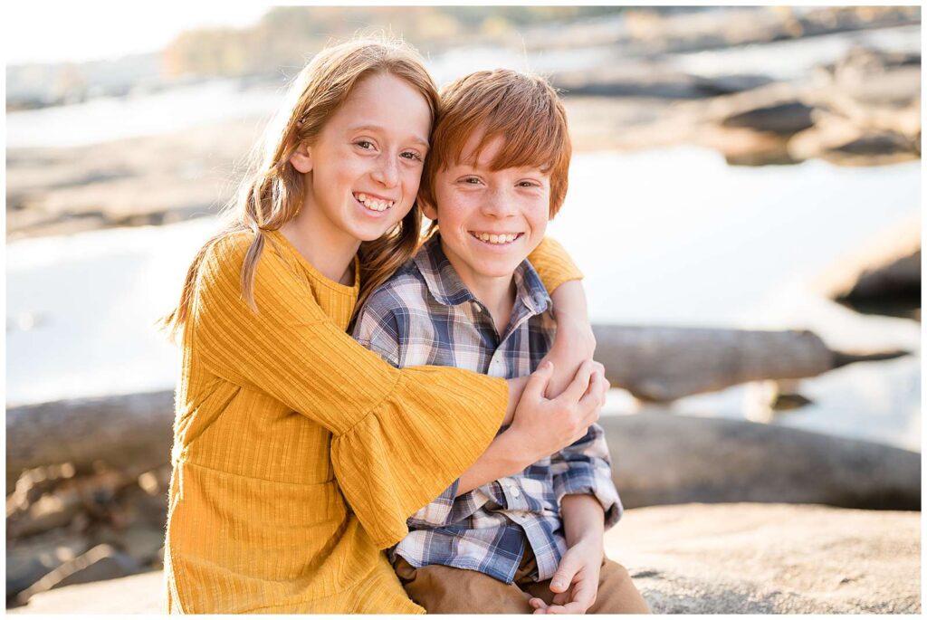Red-headed siblings-sister and brother-sit on a rock and smile at the camera of Wisp + Willow Photography Co. as sister hugs her brother.