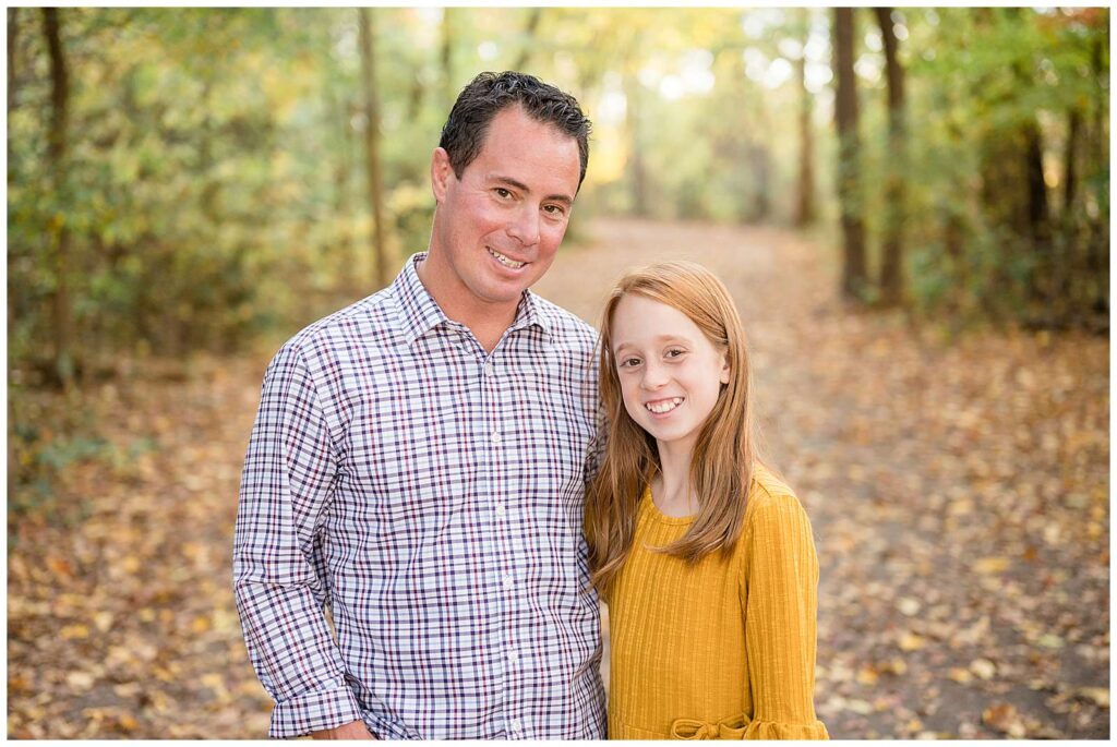Father and daughter smile at the camera of Wisp + Willow Photography Co. as they stand on a leaf covered pathway lined with trees.  Dad wears a plaid, button down shirt and daughter wears a fall, yellow dress.