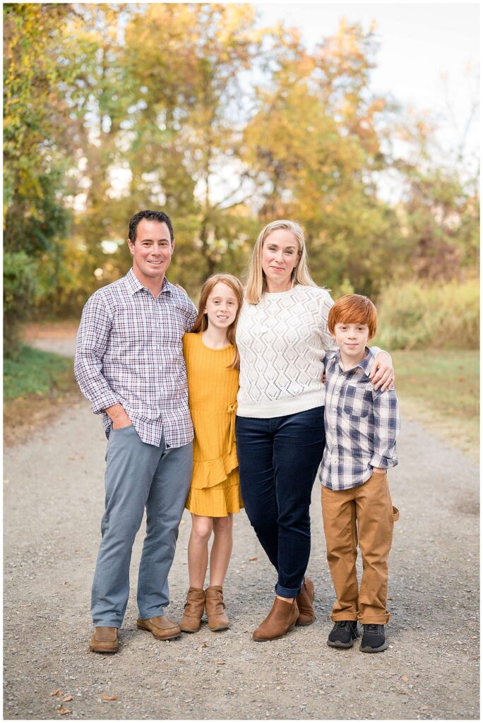 Family of 4 stand on a pathway surrounded with green grass and trees as they take a family portrait during their fall session at Belle Isle in Richmond, VA.  They wear white, blue, and yellow colors to coordinate for a fall feel.
