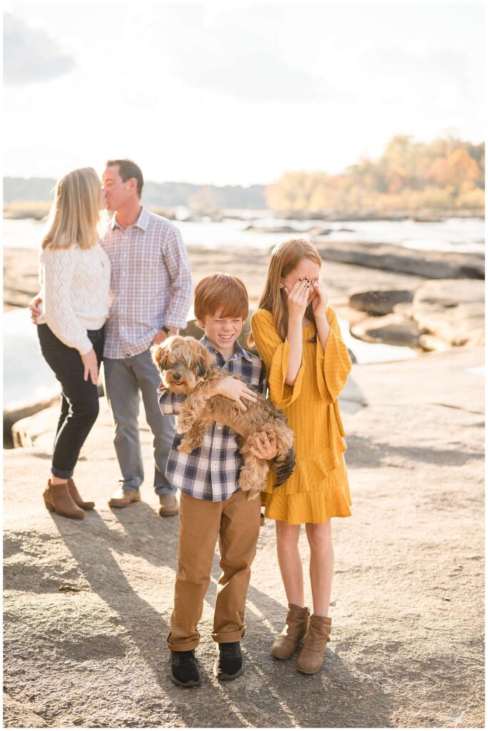 Two red-headed kids cover their eyes and make an "ew" face while their parents still a kiss behind them as they all stand on the rocks at Belle Isle in Richmond, VA!