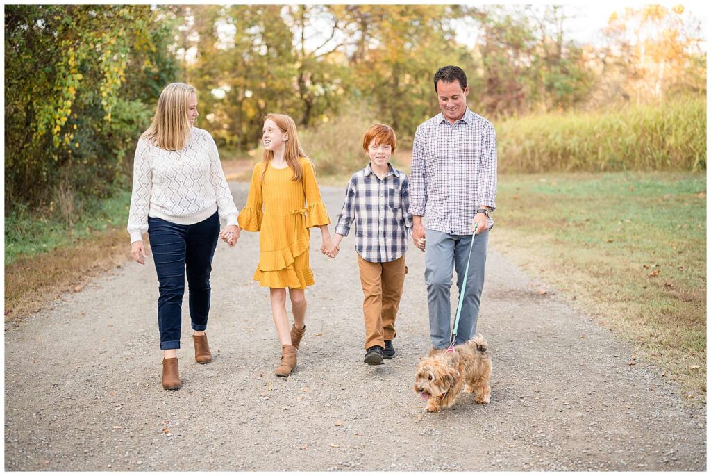 Family of 4, Mom on the left side, then daughter, son, and dad, hold hands as they walk down a pathway surrounded by green grass and trees looking at each other and their pup that Dad is walking.  Click more to see this family on the Wisp + Willow Photography Co. blog!