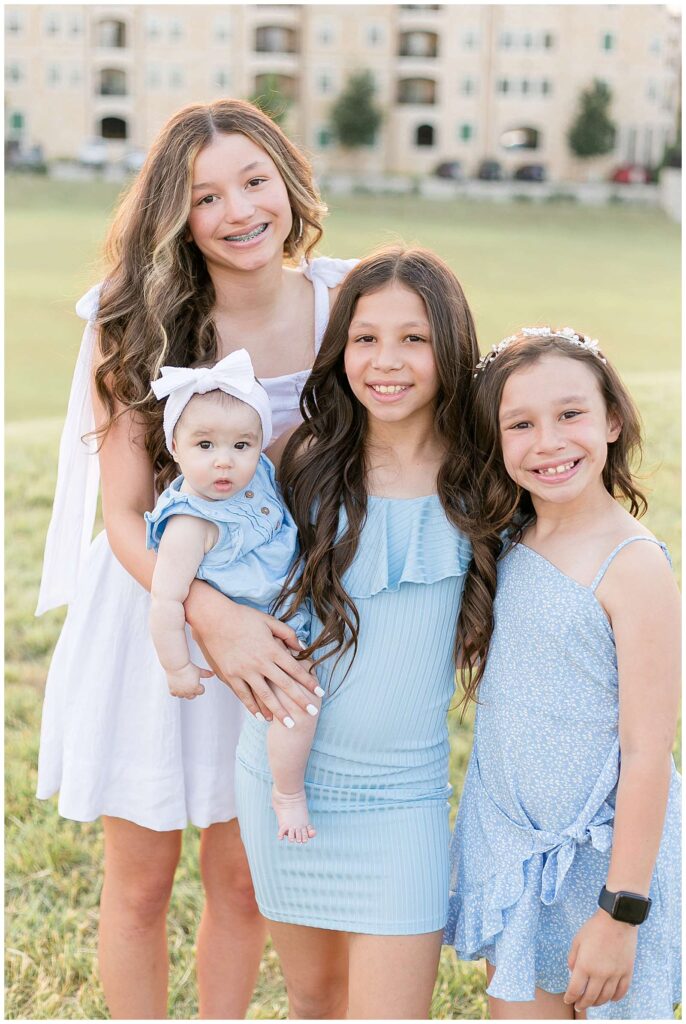 4 sisters come in close and smile at the camera during their family session at Adriatica Village in TX.  They wear coordinating dresses with white and light blue. 
