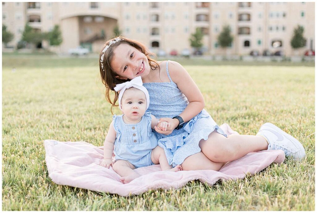 Older sister sits with her baby sister who is sitting up on a field of grass at Adriatica Village in McKinney, TX for their family portrait session.