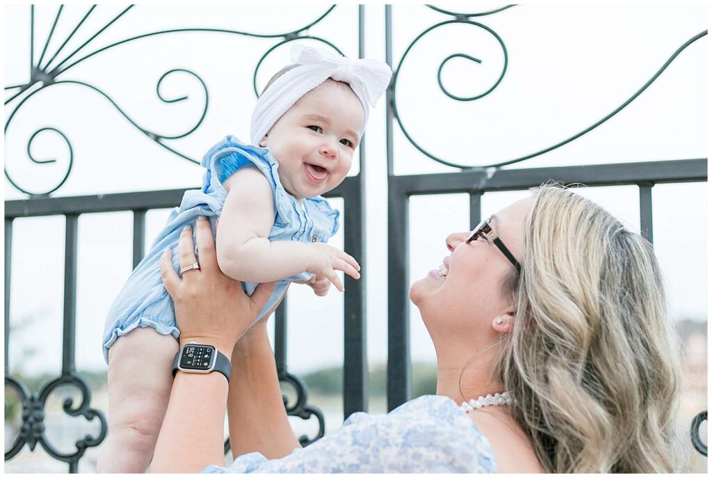 Mom, looking up and smiling, holds up her baby girl who is laughing at the camera and wears a white bow headband and a blue onesie romper during their family session in McKinney, TX.