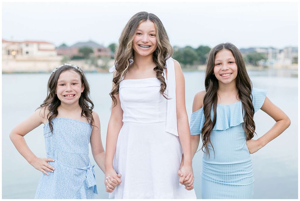 McKinney, TX photographer, Wisp + Willow Photography Co. takes a picture of 3 sisters who hold hands and smile at the camera wearing coordinate dresses of white and light blue.