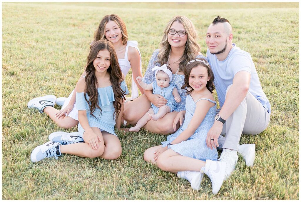 McKinney family pictures are taken of a family of 6, parents and 4 daughters, in a grass field at Adriatica Village.  They sit on the field and look up at the camera as they wear coordinating light blue outfits during their spring family session.