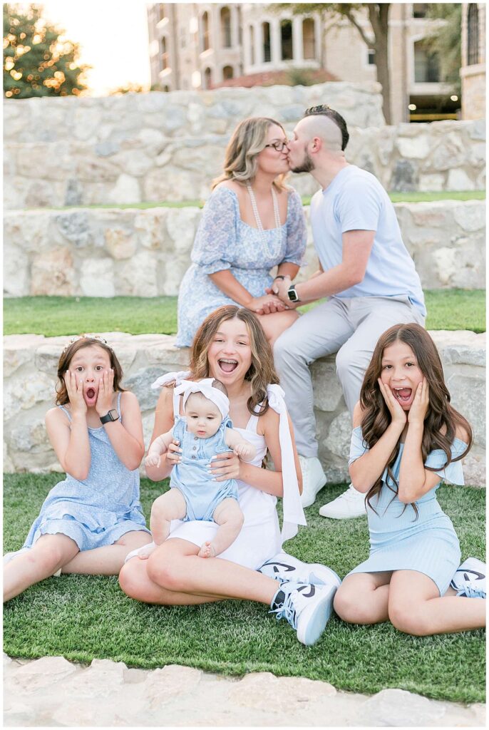 Mom and Dad kiss in the background as their 4 daughters look at the camera with faces that they can't believe they are kissing:-)  They coordinate wearing blue and white for their family session ini McKinney, TX at Adriatica Village.