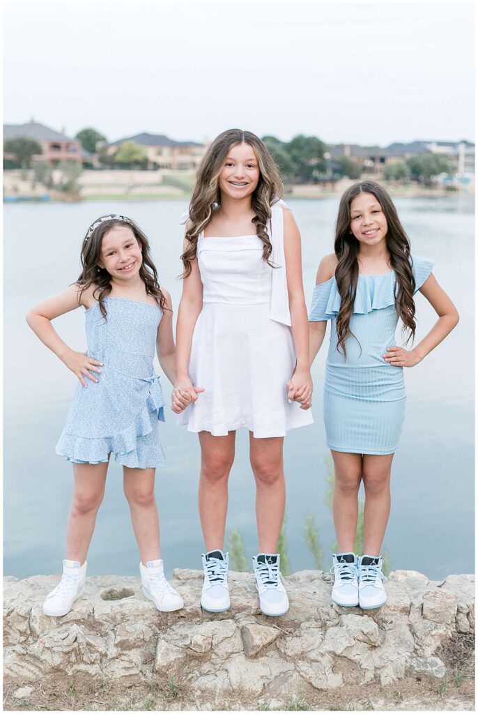 3 sisters standing in front of the water at Adriatica Village in McKinney, TX.  They are standing holding hands, and the two on the hands puts their hands on their hips.  They coordinate in light blue and white, all 3 wearing dresses.  Click to see more of this family session on the Wisp + Willow Photography Co. blog!