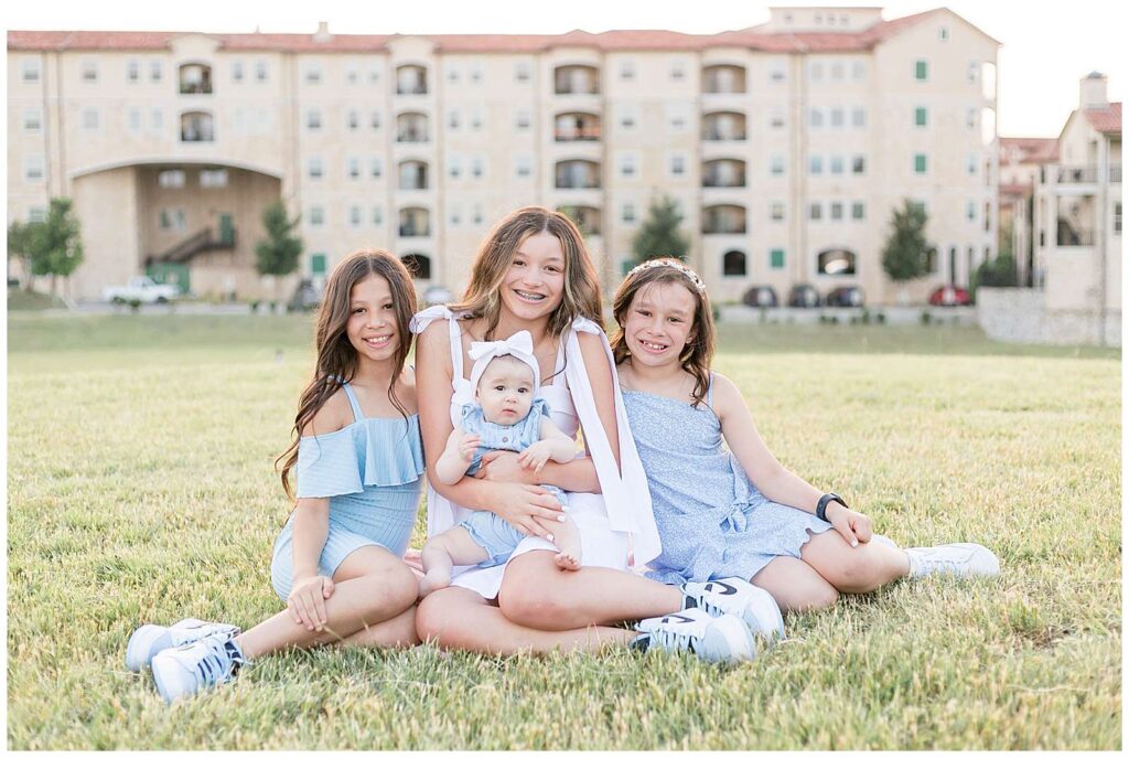 4 sisters sit on a grass field in front of a building at Adriatica Village in McKinney, TX.  They wear coordinating light blue and white for springtime during their family session with Wisp + Willow Photography Co.