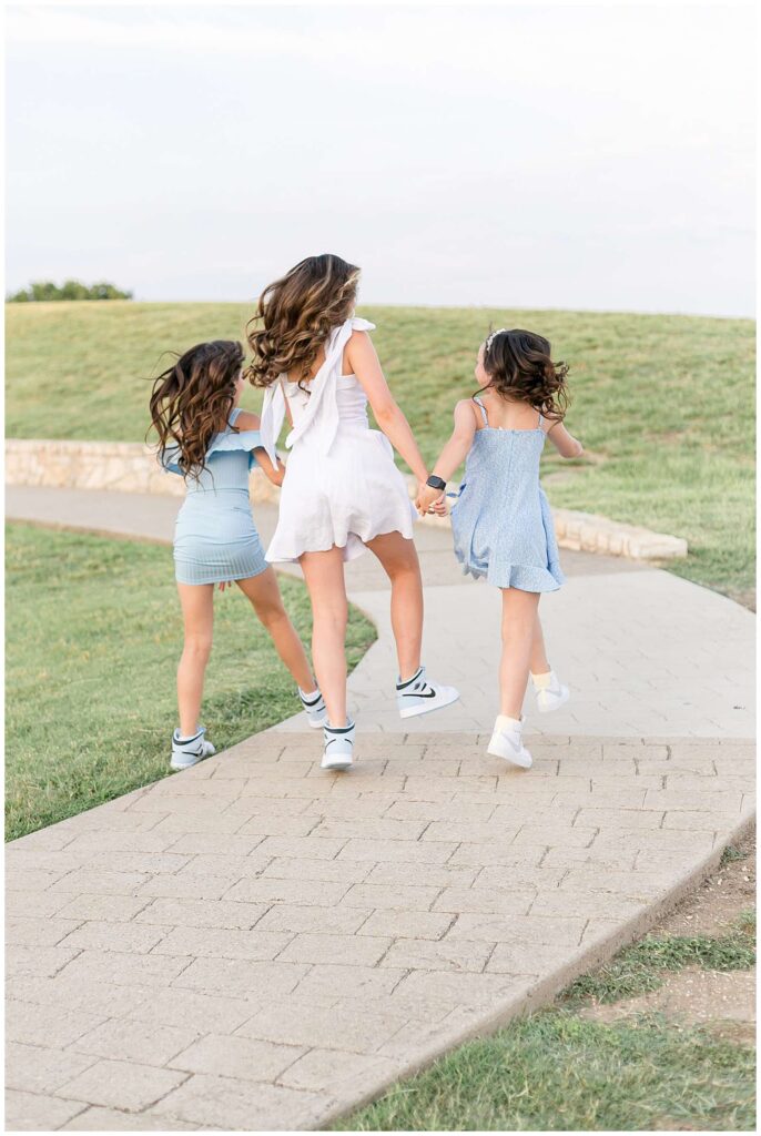 3 sisters hold hands and run away from the camera up the pathway at Adriatica Village in McKinney, TX wearing light blue and white dresses.