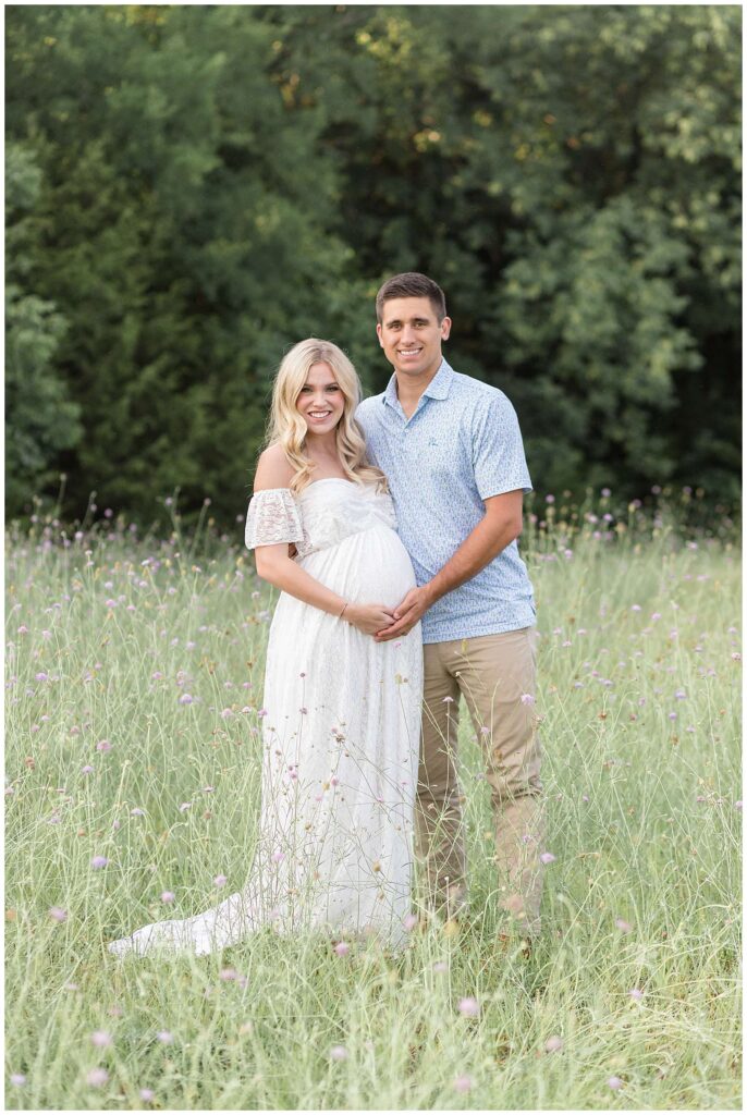 Expecting mom-to-be and husband, stand in a field with tall purple flowers as he holds is wife's belly with her and they smile at the camera of Wisp + Willow Photography Co.  Click to see more of this couple on the blog!