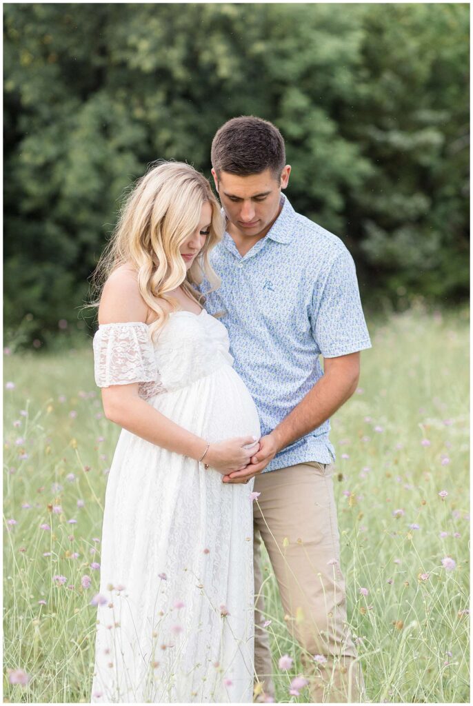 Expecting parents, wife wears a beautiful white, lace, off the the shoulders dress and her husband wears a blue, detailed shirt khakis.  He holds her belly with her and they both look down at her belly.
