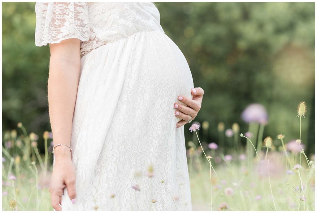 Beautiful maternity portrait taken by Wisp + Willow Photography focuses on a baby bump that is in a white, lace dress and surrounded by a field of purple wildflowers.